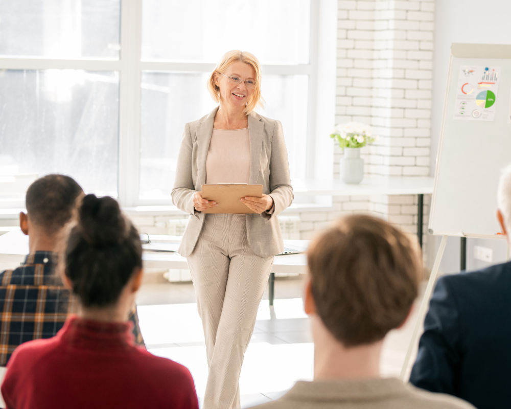 A smiling woman in a beige suit holds a tablet while speaking to a seated audience in a bright, modern room. A flipchart with colorful charts is visible in the background.