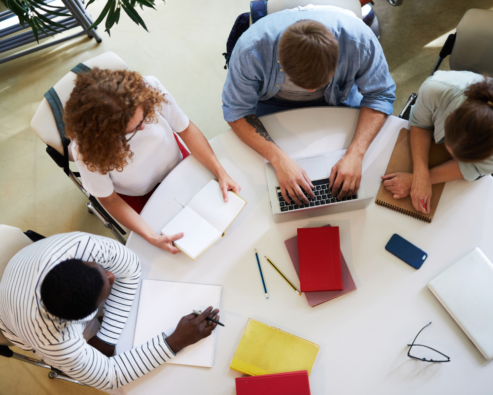 Four people sit around a table working; one uses a laptop, others write in notebooks. Books, pens, and a smartphone are scattered on the white table. The scene is viewed from above.