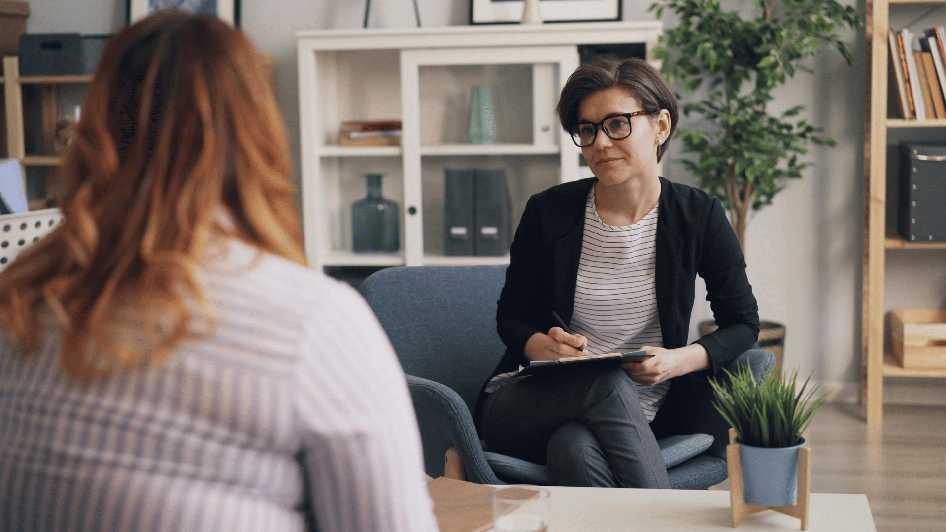 A person with short hair and glasses sits in a chair, holding a clipboard and pen, listening attentively to another person with long hair during a conversation in an office setting.