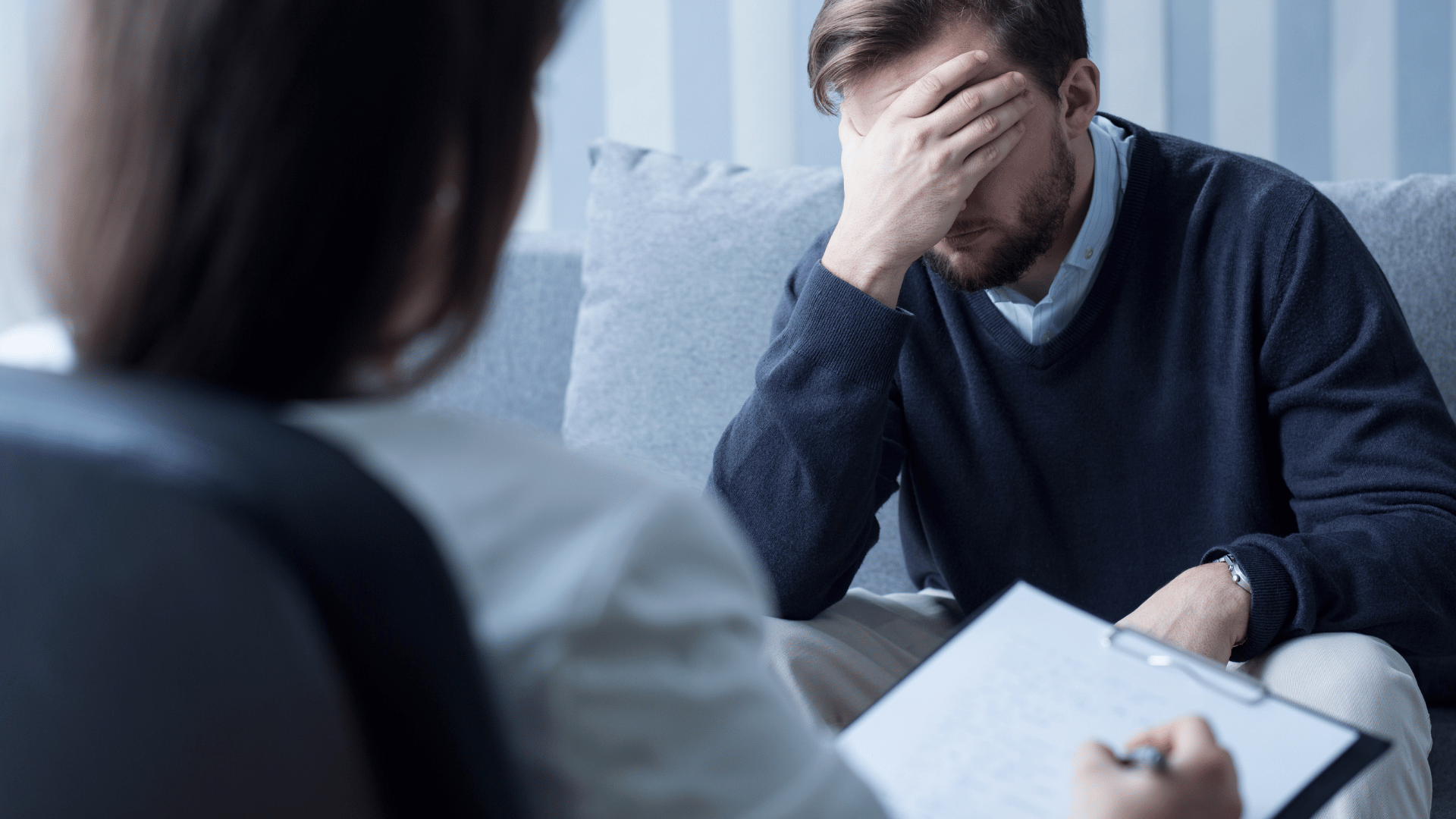A man sits on a couch with his head in his hand, appearing distressed, while another person, possibly a therapist, takes notes on a clipboard during a counseling session.