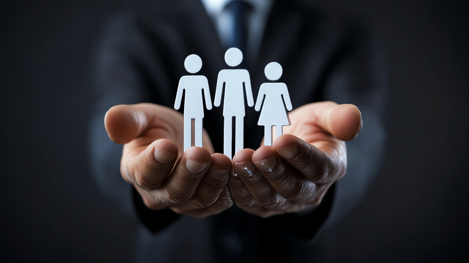 A person in a suit holds out their hands, displaying three paper cutouts of human figures, including two with pants and one with a dress, symbolizing teamwork or diversity.